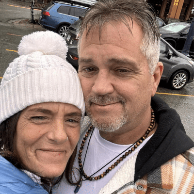 A smiling man and woman take a selfie outdoors. The woman wears a white knitted hat, and the man is in a plaid jacket with a beaded necklace. Cars and a building are in the background.
