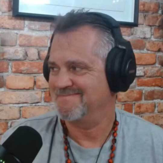 A man with headphones and a beard is smiling, sitting in front of a microphone against a brick wall background.