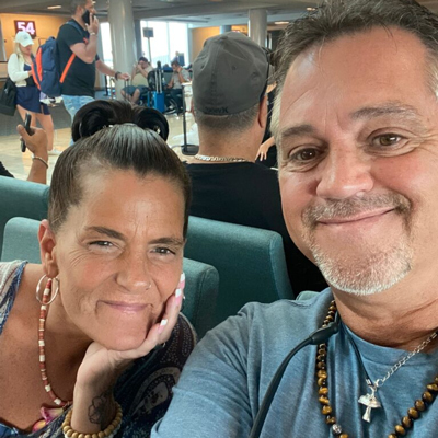 Two people smiling at the camera, seated in an airport. People are visible in the background, with some wearing backpacks. Signs and airport seating are seen in the vicinity.