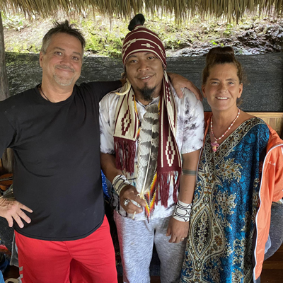 Three people standing together under a thatched roof. The person in the center is wearing a patterned scarf and accessories. The other two are dressed casually.