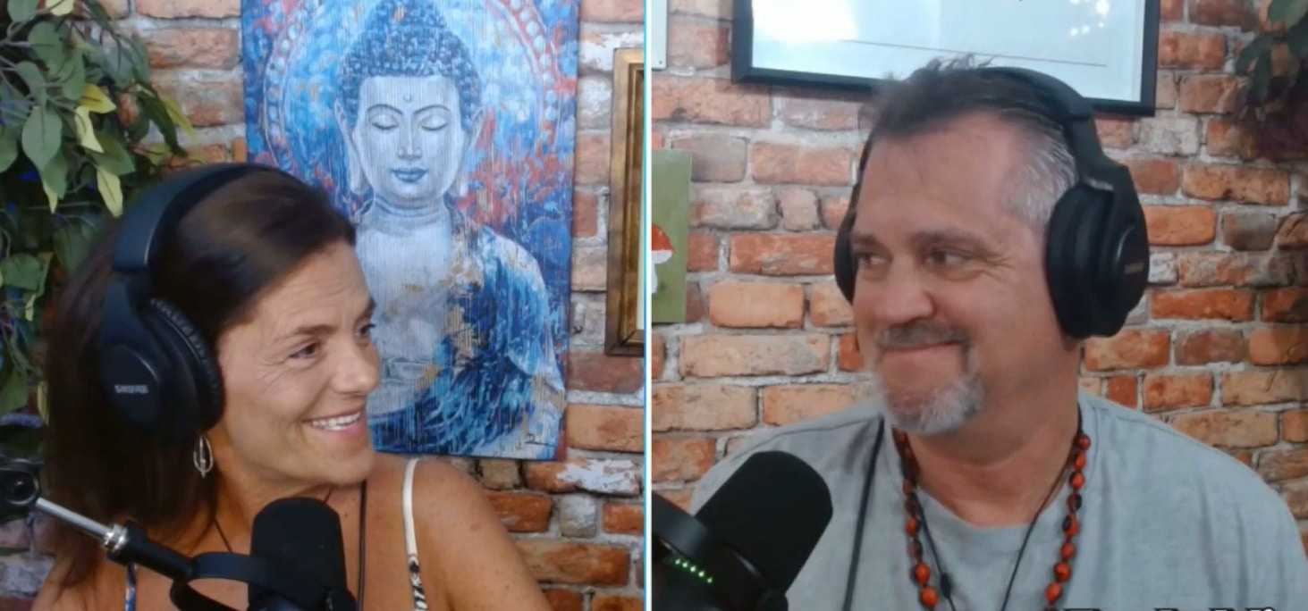 Two people wearing headphones are smiling at each other while speaking into microphones. A Buddha painting is on the brick wall behind them.