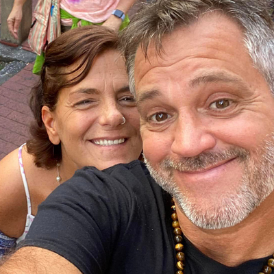 Two people taking a selfie outdoors. The person in front has short hair and a beard, wearing a black shirt and necklace. The person behind has shoulder-length hair. Both are smiling.