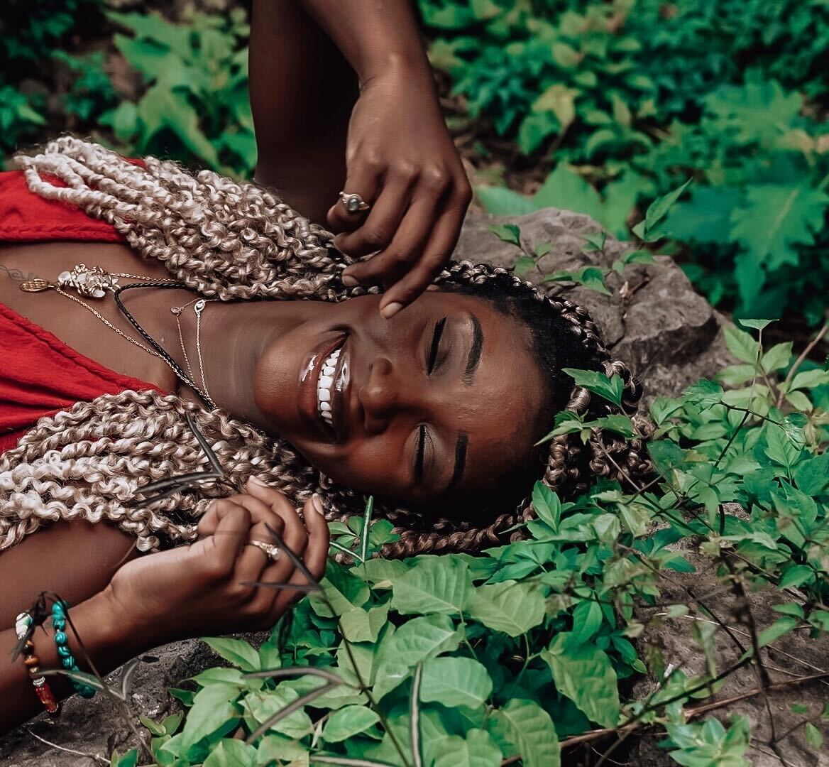Person lying on their back outdoors, surrounded by green plants, with eyes closed and smiling. They're wearing a red top, necklaces, and bracelets.