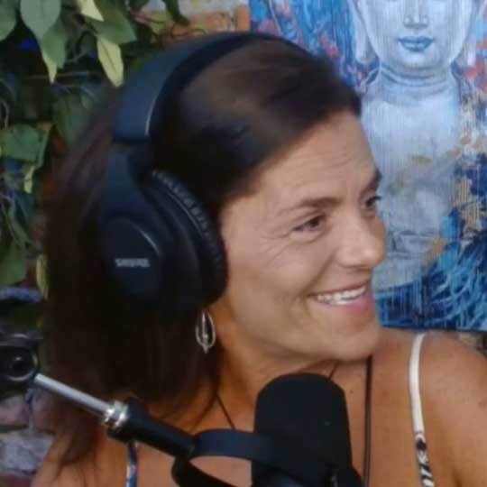 A woman with long brown hair wearing large headphones sits in front of a microphone, smiling brightly, as she discusses microdosing. Behind her, a colorful painting and green leaves add vibrancy to the scene.