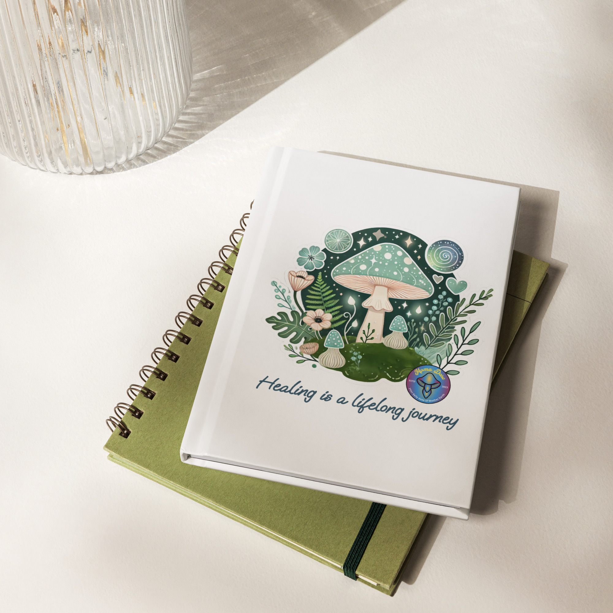 The Healing Journey Journal, a white hardcover featuring a mushroom and floral design with the quote "Healing is a lifelong journey," sits atop a green spiral notebook on a white surface.