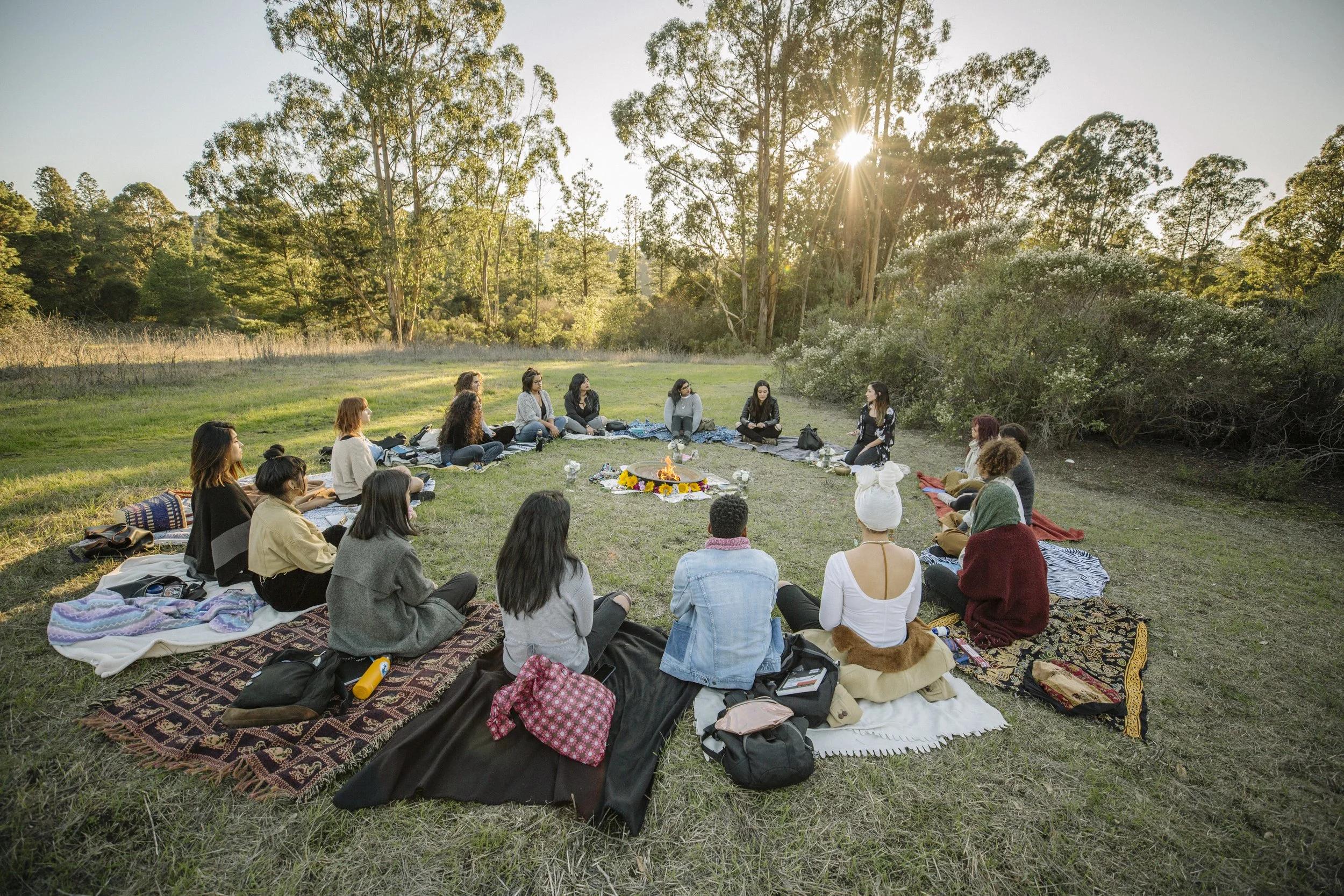 Group of people seated in a wide circle on blankets around a small fire in a clearing at sunset