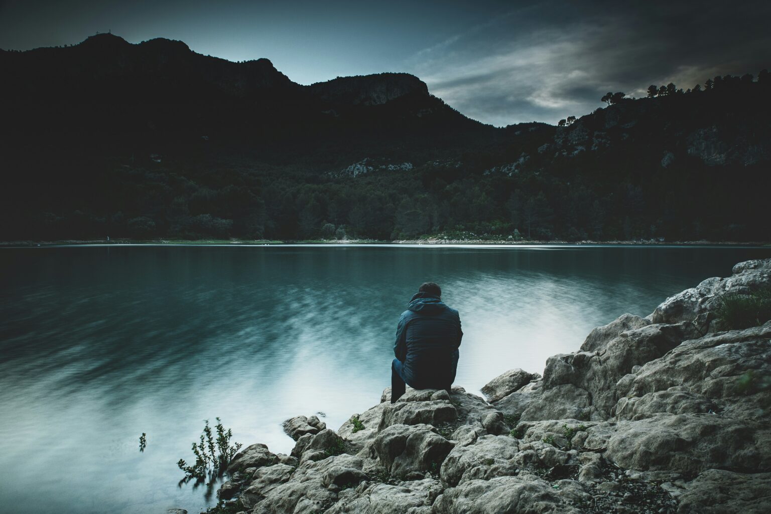 Person sitting on a rock beside a lake at dusk, facing distant mountains. healing-vs-feeling-good-lake-reflection