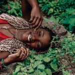 Person lying on their back outdoors, surrounded by green plants, with eyes closed and smiling. They're wearing a red top, necklaces, and bracelets.