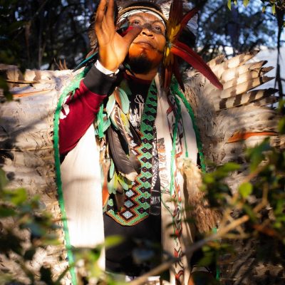 A person wearing a traditional feathered headdress and colorful garments stands outdoors, partially surrounded by foliage, with one hand raised to their face.