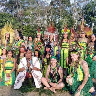 A group of people in vibrant traditional attire pose outdoors, surrounded by greenery and trees.