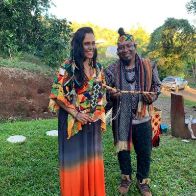 Two people in colorful traditional attire stand on grass outdoors, smiling and holding a decorative stick, with trees and a parked car in the background.