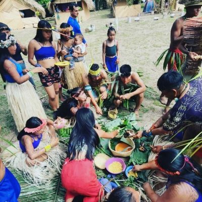 A group of people in traditional attire prepare food on large leaves outdoors. Some are sitting, others are standing, and a child is visible in the scene.