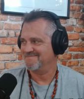 Man with a beard smiling, wearing headphones and a necklace, sitting in front of a brick wall with a microphone.