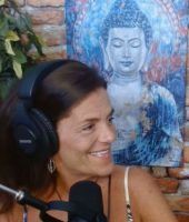 Person wearing headphones speaking into a microphone, with a Buddha painting on a brick wall backdrop.